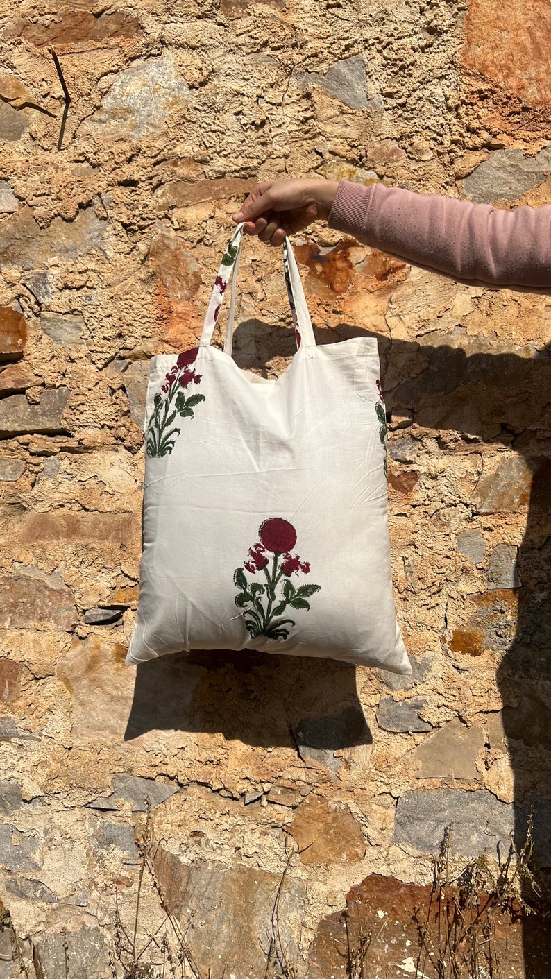 May include: White cotton tote bag with red and green floral print. The bag has two handles and is held by a hand.