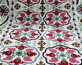 May include: White textile with intricate embroidery featuring floral and geometric patterns in red, pink, blue, green, and brown. The design includes stylized flowers and symmetrical arrangements, creating a detailed and colorful textile.