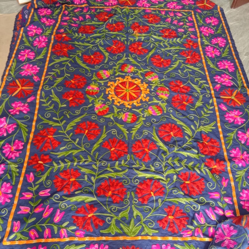 May include: A blue fabric tapestry with intricate embroidery featuring red, pink, and yellow flowers. The design is symmetrical and includes a central circular motif.