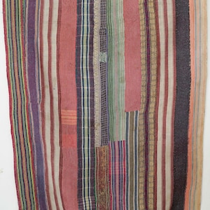 May include: A patchwork textile with vertical stripes in various colors, including red, purple, and beige. The textile has a rectangular shape and appears to be a quilt or blanket. The edges are finished with a border.