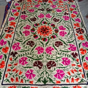 May include: A white cotton fabric with intricate embroidery in shades of pink, orange, green, and brown. The design features a central floral motif surrounded by smaller floral patterns.