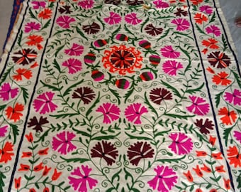 May include: A white cotton fabric with intricate embroidery in shades of pink, orange, green, and brown. The design features a central floral motif surrounded by smaller floral patterns.