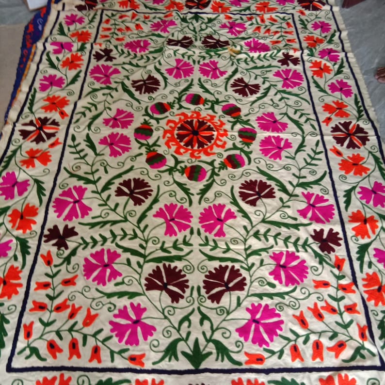 May include: A white cotton fabric with intricate embroidery in shades of pink, orange, green, and brown. The design features a central floral motif surrounded by smaller floral patterns.