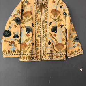 May include: A beige embroidered jacket with a floral pattern in shades of brown, blue, and green. The jacket has a mandarin collar and a zipper closure.