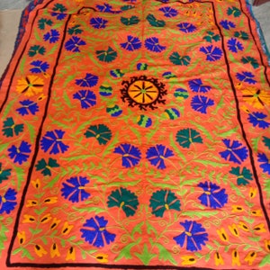 May include: A large, rectangular, orange fabric with intricate embroidery. The embroidery features a central design of a wheel with radiating spokes, surrounded by stylized flowers in shades of blue, green, and yellow.