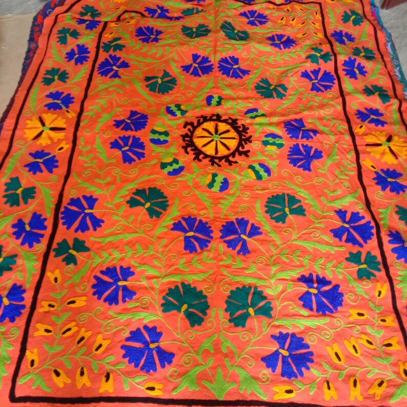 May include: A large, rectangular, orange fabric with intricate embroidery. The embroidery features a central design of a wheel with radiating spokes, surrounded by stylized flowers in shades of blue, green, and yellow.