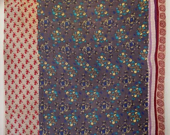 Op de afbeelding: Een rechthoekige textielquilt met een centraal paneel van donkergrijze stof met een herhalend blauw en geel bloemenpatroon. De quilt is omrand door een crèmekleurig gedeelte met kleine rode bloemen en een verticale strook met een rode paisleyprint.