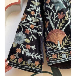 May include: Black velvet jacket with detailed floral embroidery in orange, blue, and green. The jacket has wide sleeves and decorative stitching. A stylish piece to add a bohemian touch to any outfit. The jacket's measurements are in centimetres.