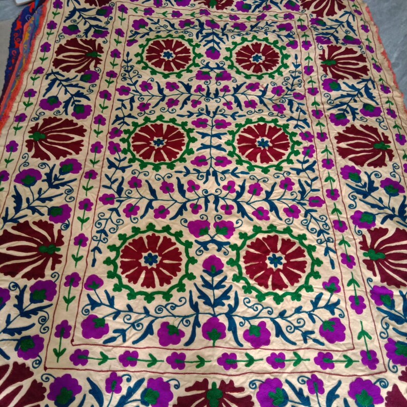May include: A cream-colored fabric with intricate embroidery in shades of red, green, blue, and purple. The design features floral motifs and geometric patterns.