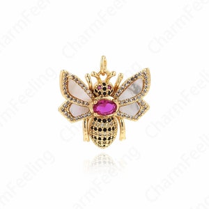 May include: A gold bee charm with black and white accents. The bee has a pink gem in the center of its body and white wings with black accents.