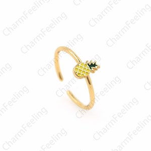 Könnte beinhalten: Ein goldfarbener, verstellbarer Ring mit einem kleinen, gelben Ananas-Anhänger mit grünen Blättern. Der Ring ist offen, um die Größe anzupassen. Der Ananas-Anhänger hat ein strukturiertes Aussehen.