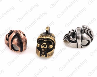 May include: Three metal skull helmet charms in different colors: copper, gold, and silver. The charms are detailed with a skull face and a helmet design.