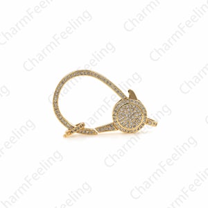 May include: A gold-toned metal clasp with a round, clear crystal-encrusted design. The clasp is designed to attach to a chain or bracelet.