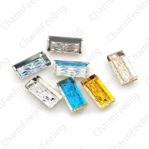 Peut inclure: Assortiment de huit strass rectangulaires en verre de différentes couleurs, notamment transparent, bleu, jaune et gris. Les strass ont un design facetté et sont éparpillés sur une surface blanche.