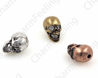 May include: Three skull-shaped beads with rhinestone eyes. The beads are silver, gold, and copper in color.