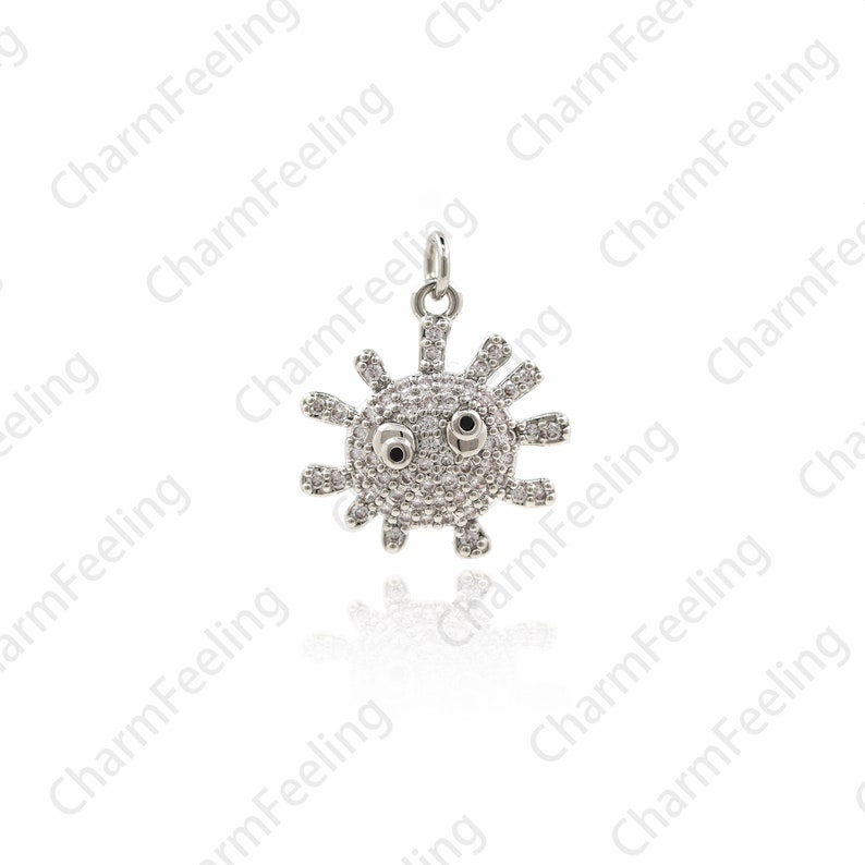 May include: A silver charm in the shape of a sun with a face. The sun has small, round, white stones for eyes and a black center. The sun is surrounded by a ring of small, white stones.