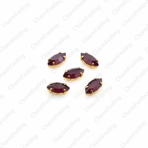May include: Six maroon marquise-shaped rhinestones with gold settings. The rhinestones are scattered on a white background.