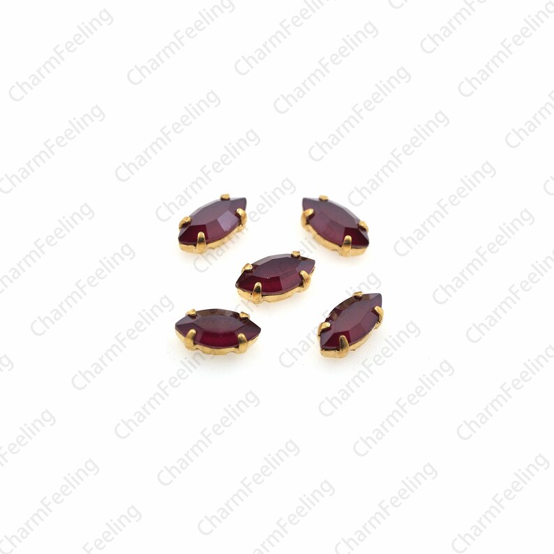 May include: Six maroon marquise-shaped rhinestones with gold settings. The rhinestones are scattered on a white background.