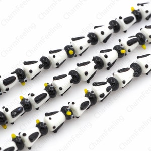 May include: Strands of glass beads shaped like cows. Each bead is white with black spots and a black head. The cows have yellow horns. The beads are strung together, creating a playful and unique design.