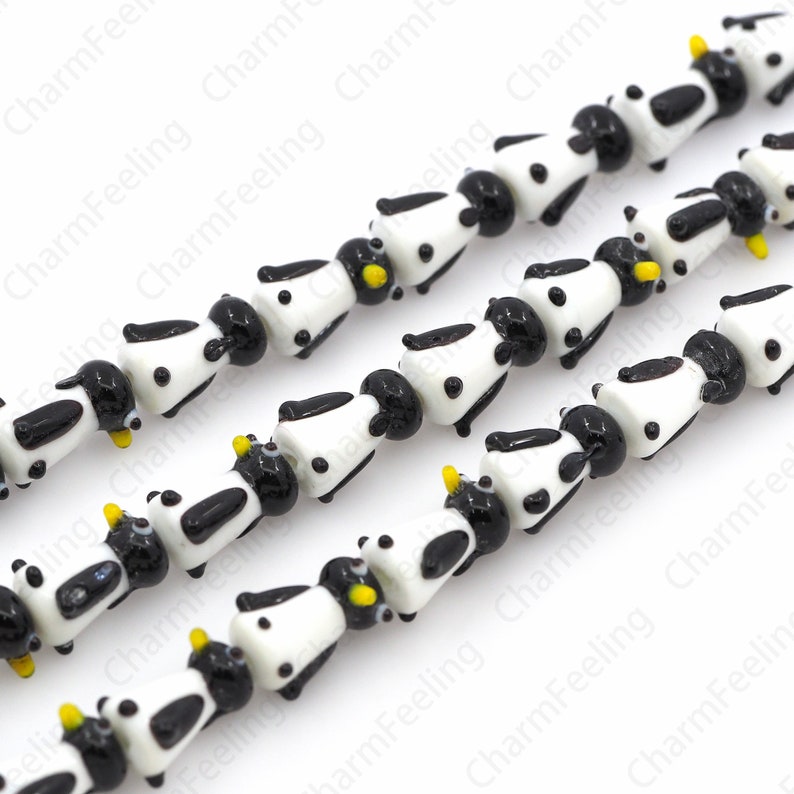 May include: Strands of glass beads shaped like cows. Each bead is white with black spots and a black head. The cows have yellow horns. The beads are strung together, creating a playful and unique design.