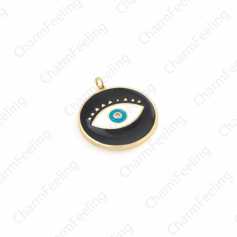K&ouml;nnte beinhalten: Ein goldfarbener Anh&auml;nger mit einem schwarzen Emaille-Kreis, der ein wei&szlig;es Auge mit einer blauen Pupille und einem kleinen, klaren Strassstein in der Mitte zeigt. Das Auge ist von einer Reihe kleiner Dreiecke umgeben.