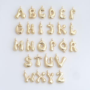 May include: A set of gold-coloured alphabet charms, each with a rounded, bubble-like design. The letters A to Z are displayed, each with a small loop at the top for attaching to a chain or bracelet. The charms are arranged in neat rows.