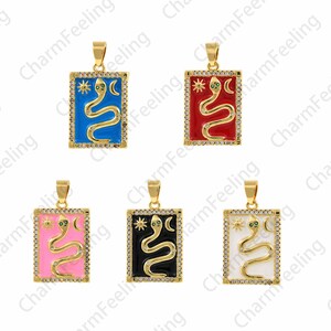 Op de afbeelding: Vijf gouden hangers met een slangenontwerp. De hangers zijn rechthoekig en hebben een witte rand met kleine, heldere stenen. Elke hanger heeft een anders gekleurde emaille achtergrond: blauw, rood, roze, zwart en wit.