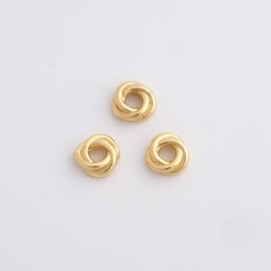 Puede incluir: Tres cuentas doradas entrelazadas en forma de nudo están dispuestas sobre una superficie blanca. Cada cuenta tiene un acabado liso y pulido y un diseño circular, lo que sugiere componentes de joyería o elementos decorativos.