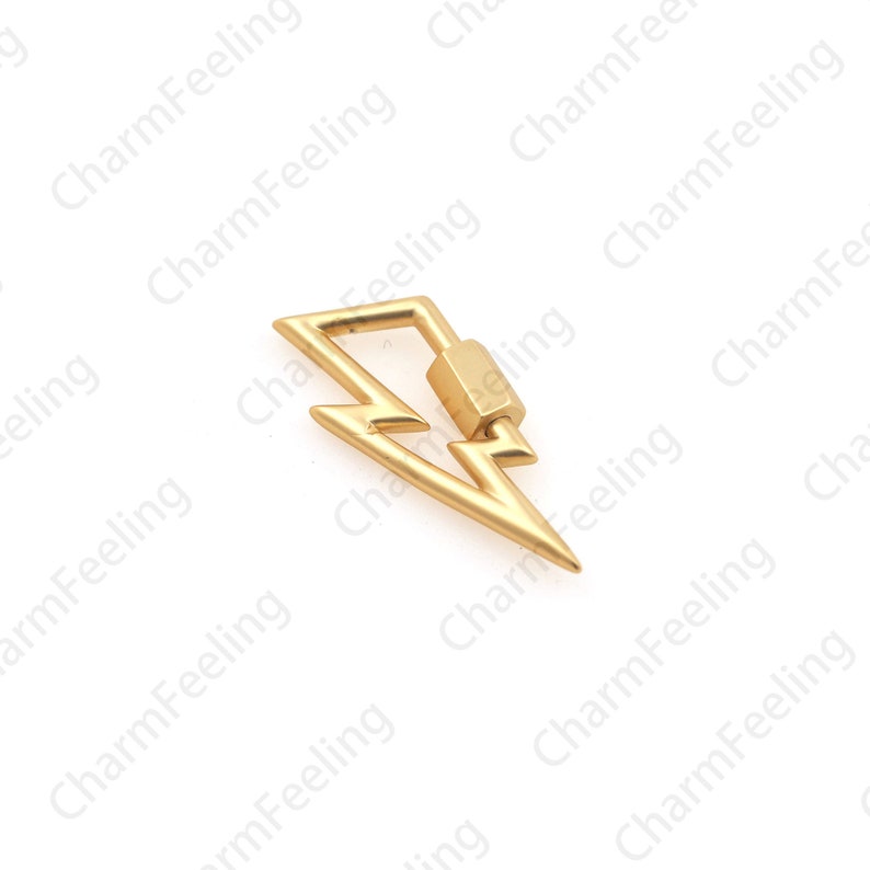 May include: A gold-colored lightning bolt charm with a rectangular clasp. The charm is a stylized representation of a lightning bolt, with sharp angles and a pointed end. The background is white.