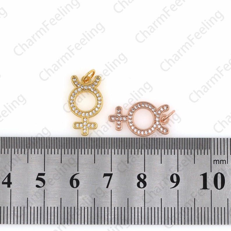 May include: Two gold and rose gold charms in the shape of the female symbol with a circle around it. The charms are decorated with clear rhinestones. The charms are measured against a ruler with markings from 4 to 10 centimeters.