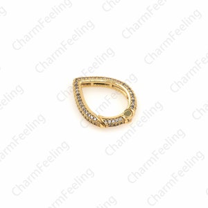May include: A gold-toned teardrop-shaped charm with clear cubic zirconia stones. The charm has a hinged opening, allowing it to be attached to a bracelet or necklace.