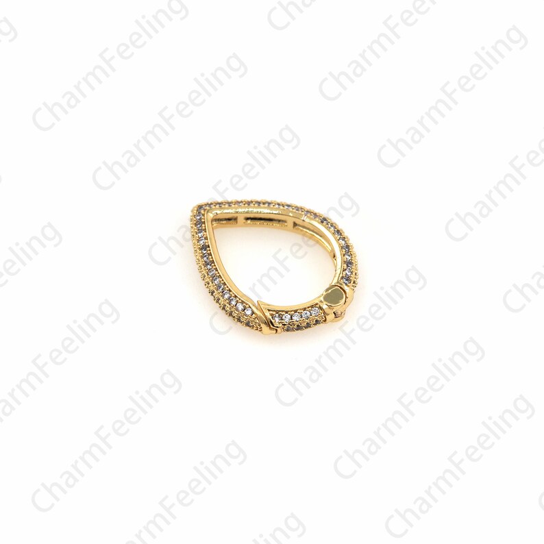 May include: A gold-toned teardrop-shaped charm with clear cubic zirconia stones. The charm has a hinged opening, allowing it to be attached to a bracelet or necklace.