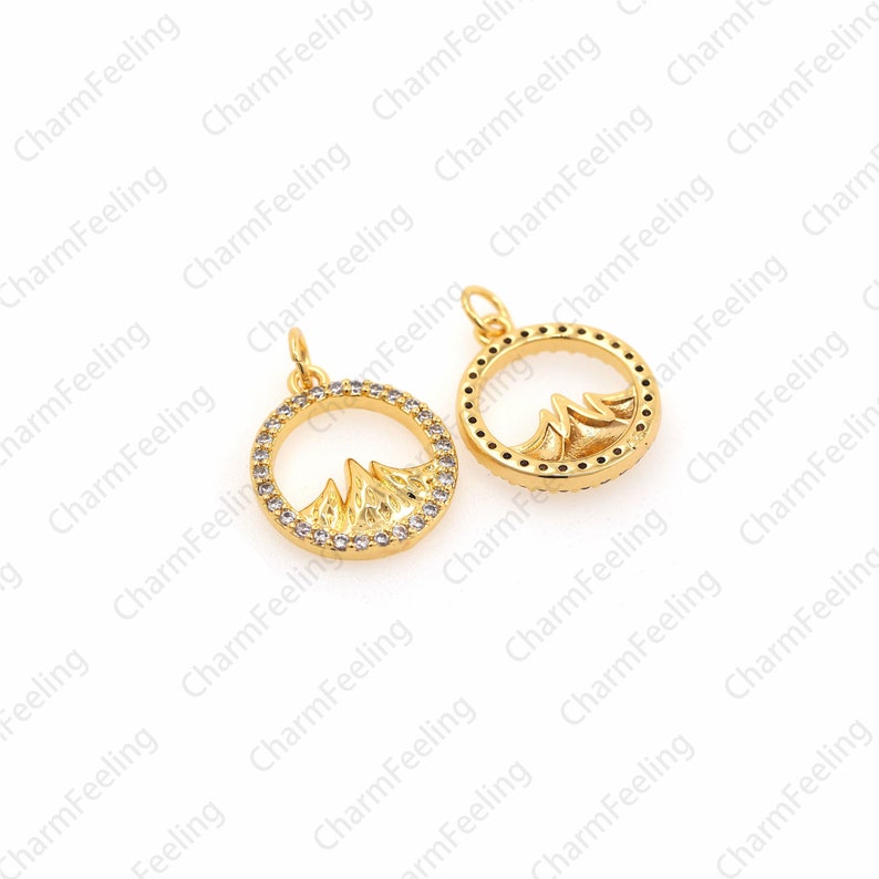 May include: Two gold-tone pendant charms featuring a mountain range design within a circle. The circles are outlined with clear rhinestones.