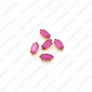 May include: Five pink glass marquise-shaped rhinestones with gold settings. The rhinestones are scattered on a white background.