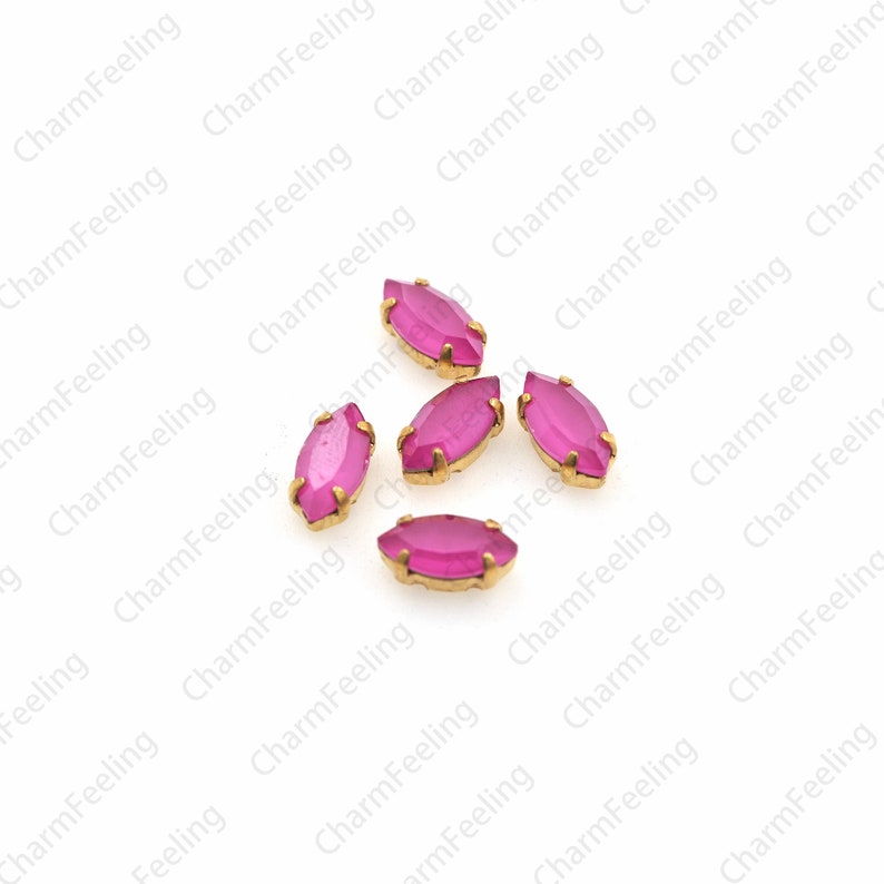 May include: Five pink glass marquise-shaped rhinestones with gold settings. The rhinestones are scattered on a white background.