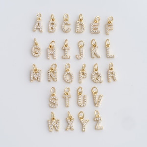 May include: A collection of gold-colored alphabet charms, each encrusted with small, clear crystals. The letters A through Z are displayed, each with a small loop for attaching to a necklace or bracelet. The charms are arranged on a light background.