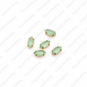 May include: Six light green marquise-shaped glass rhinestones with gold settings. The rhinestones are scattered on a white background.