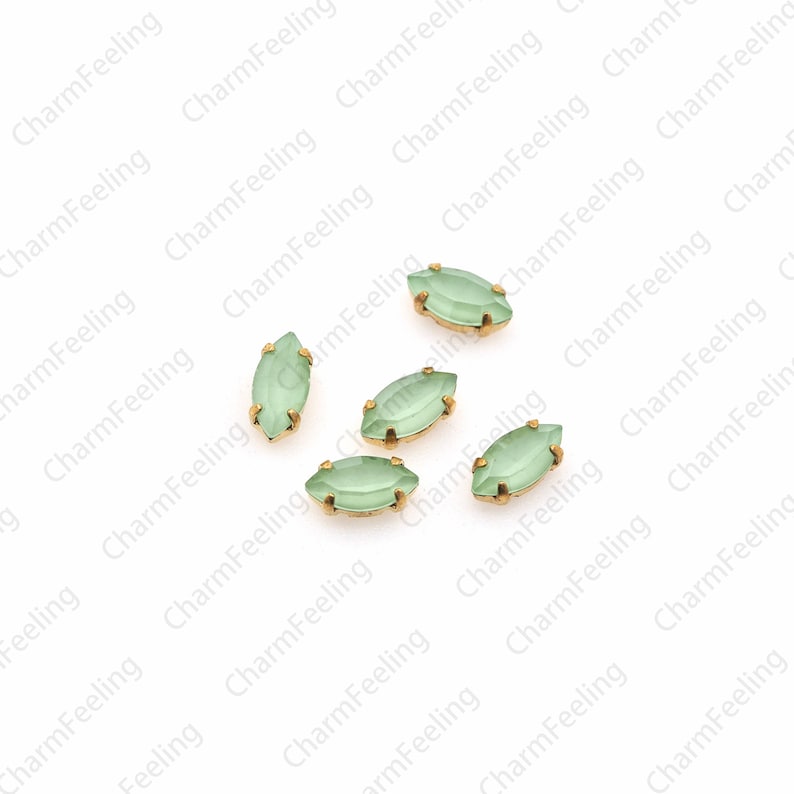 May include: Six light green marquise-shaped glass rhinestones with gold settings. The rhinestones are scattered on a white background.