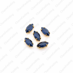 May include: Six blue marquise-shaped rhinestones with gold settings. The rhinestones are scattered on a white background.