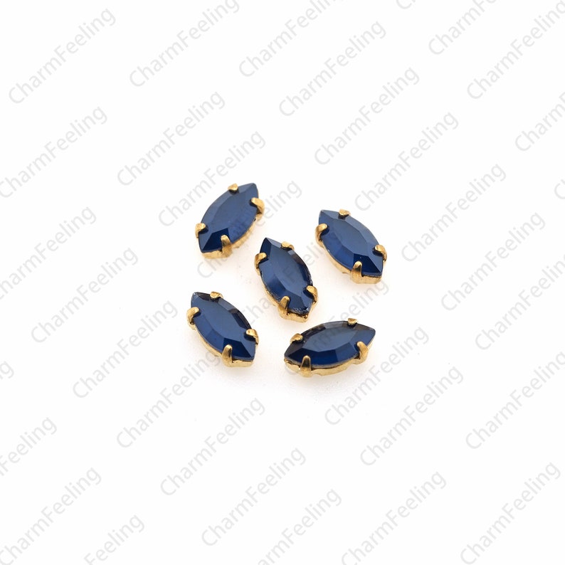 May include: Six blue marquise-shaped rhinestones with gold settings. The rhinestones are scattered on a white background.