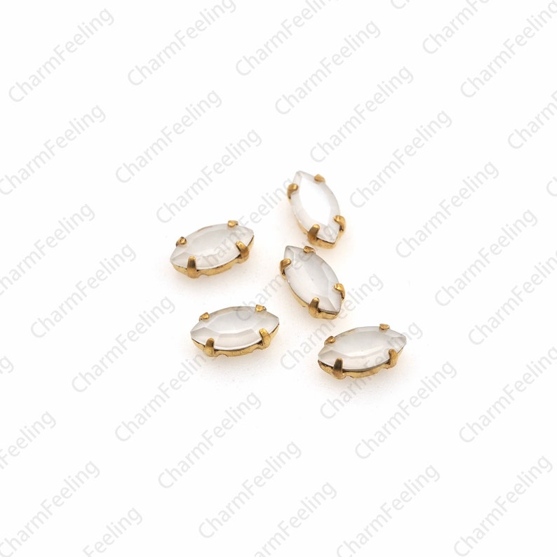 May include: Six white marquise-shaped rhinestones with gold settings. The rhinestones are scattered on a white background.