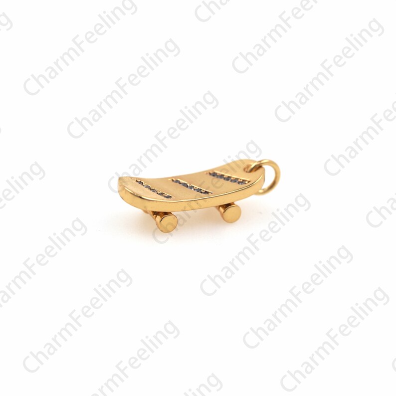 Op de afbeelding: Een gouden skateboardbedel met kleine zwarte steentjes op het dek. De bedel heeft een lus aan de bovenkant om aan een ketting of armband te bevestigen.
