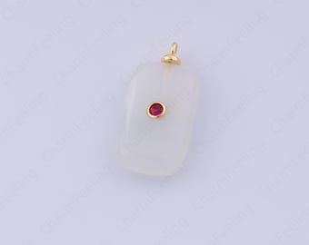 Puede incluir: Un colgante rectangular con una piedra blanca cremosa y una pequeña gema roja redonda en el centro. El colgante tiene una fianza dorada en la parte superior para sujetarlo a una cadena. El fondo es de color blanco roto.