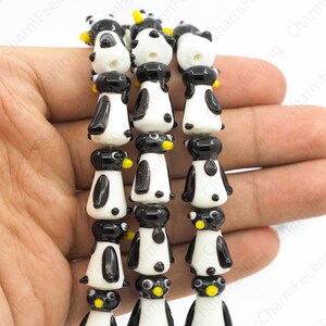 May include: Strands of glass beads shaped like penguins. Each penguin has a black head and wings, a white body, and a yellow beak. The beads are strung together, creating a playful and whimsical design, perfect for jewelry making or crafts.