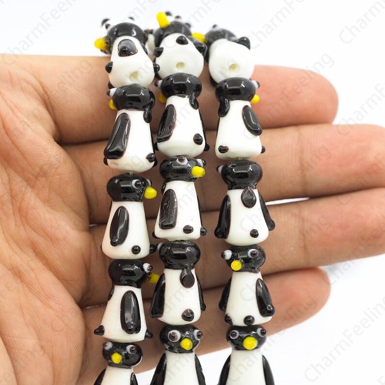 May include: Strands of glass beads shaped like penguins. Each penguin has a black head and wings, a white body, and a yellow beak. The beads are strung together, creating a playful and whimsical design, perfect for jewelry making or crafts.