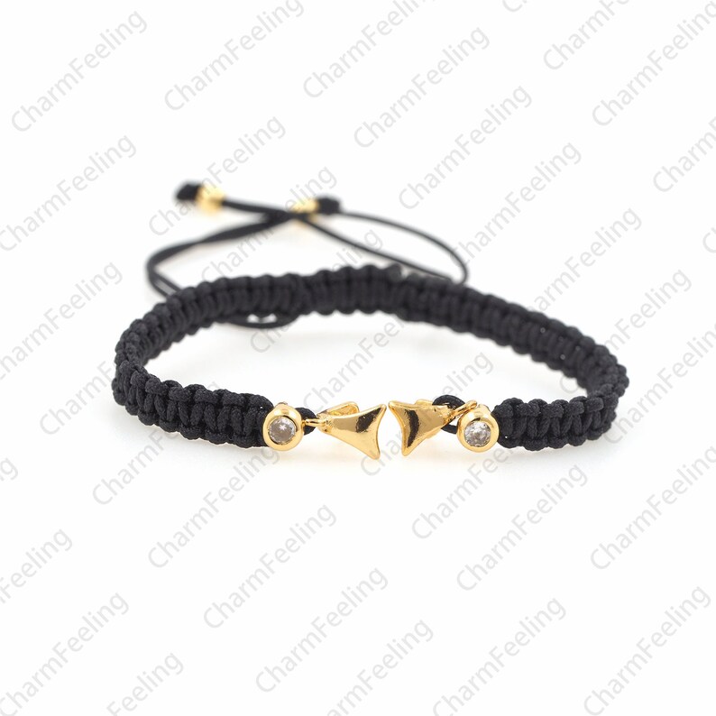 Puede incluir: Un brazalete trenzado negro con dijes de flecha dorada y detalles de cristal transparente.