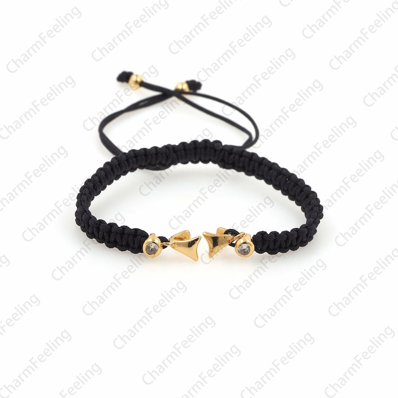 Puede incluir: Un brazalete trenzado negro con un dije dorado que presenta dos puntas de flecha con diamantes de imitaci&oacute;n transparentes. El brazalete tiene un cord&oacute;n negro que se puede ajustar para adaptarse a diferentes tama&ntilde;os de mu&ntilde;eca.