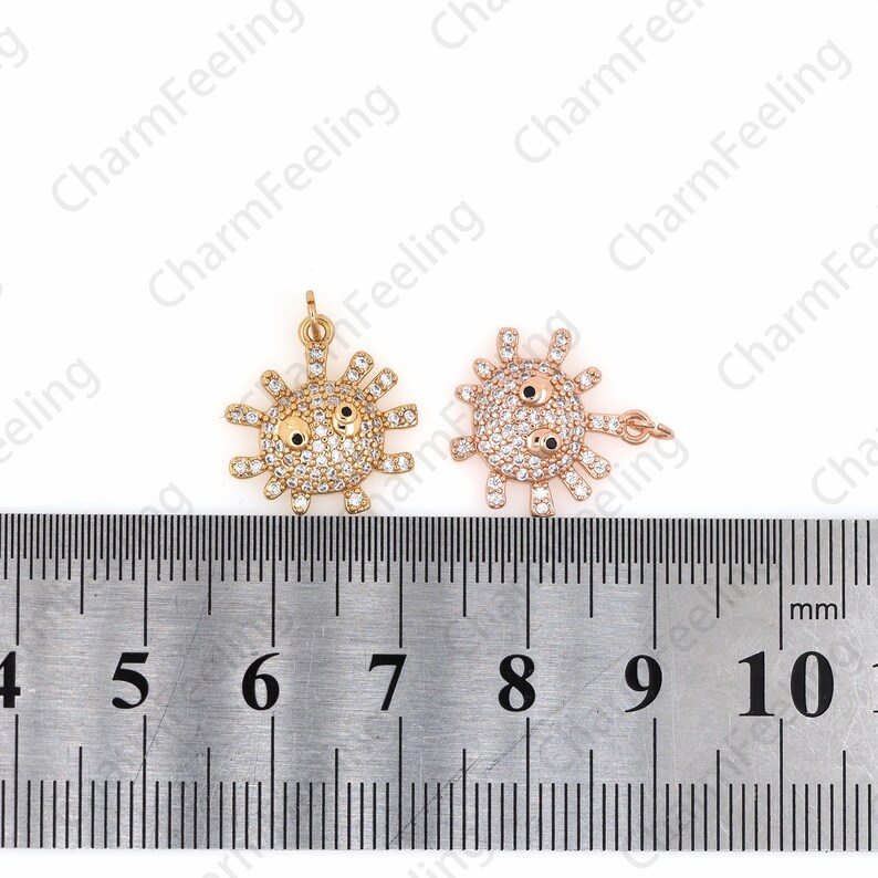 May include: Two gold-tone charms in the shape of a cartoon sun with a face. The charms are decorated with clear rhinestones and black enamel eyes. The charms are measured against a ruler, showing they are approximately 1 inch in diameter.