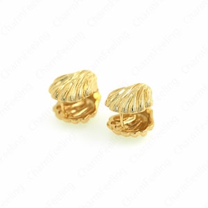 Könnte beinhalten: Zwei goldfarbene Ohrringe in Muschelform. Jeder Ohrring hat ein detailliertes, strukturiertes Design, das die natürlichen Rippen einer Muschel nachahmt. Die Ohrringe werden vor einem weißen Hintergrund präsentiert, der ihre aufwendige Handwerkskunst hervorhebt.