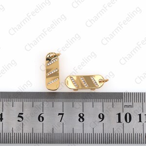 Op de afbeelding: Twee goudkleurige metalen bedels met heldere strass-accenten. De bedels worden gemeten op een liniaal met markeringen van 4 tot 11 centimeter.